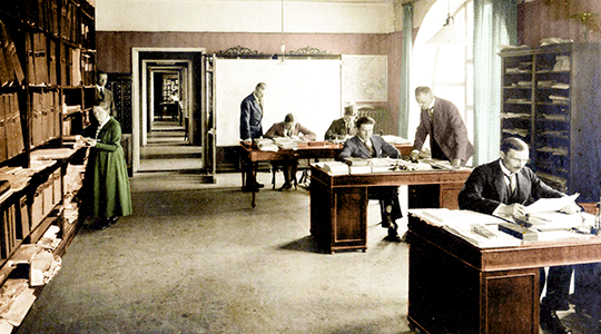 Ett gammalt fotografi där det står en kvinna vid en bokhylla, sex män sitter vid arbetsbord.